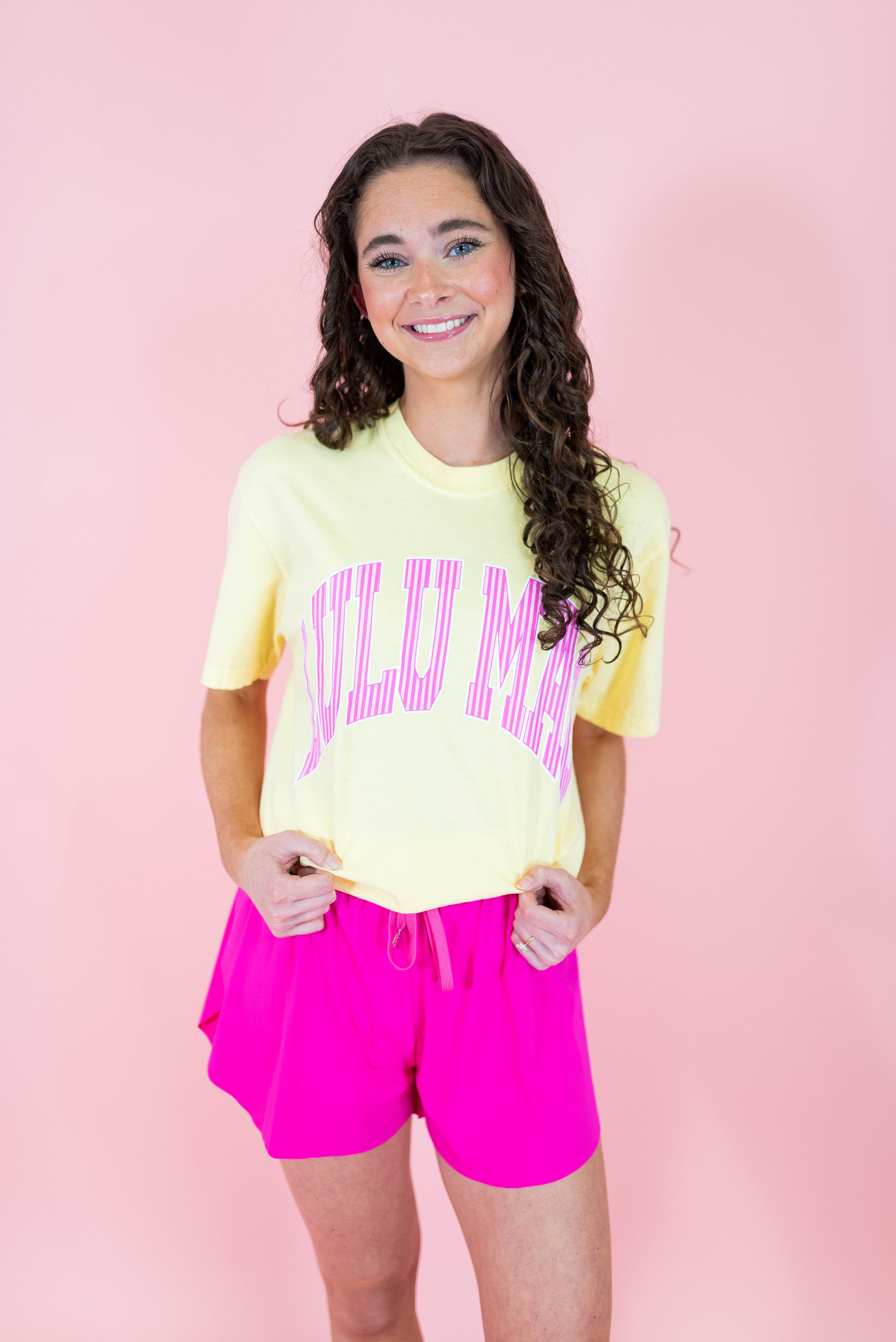Person wearing a yellow t-shirt with pink text and pink shorts on a pink background