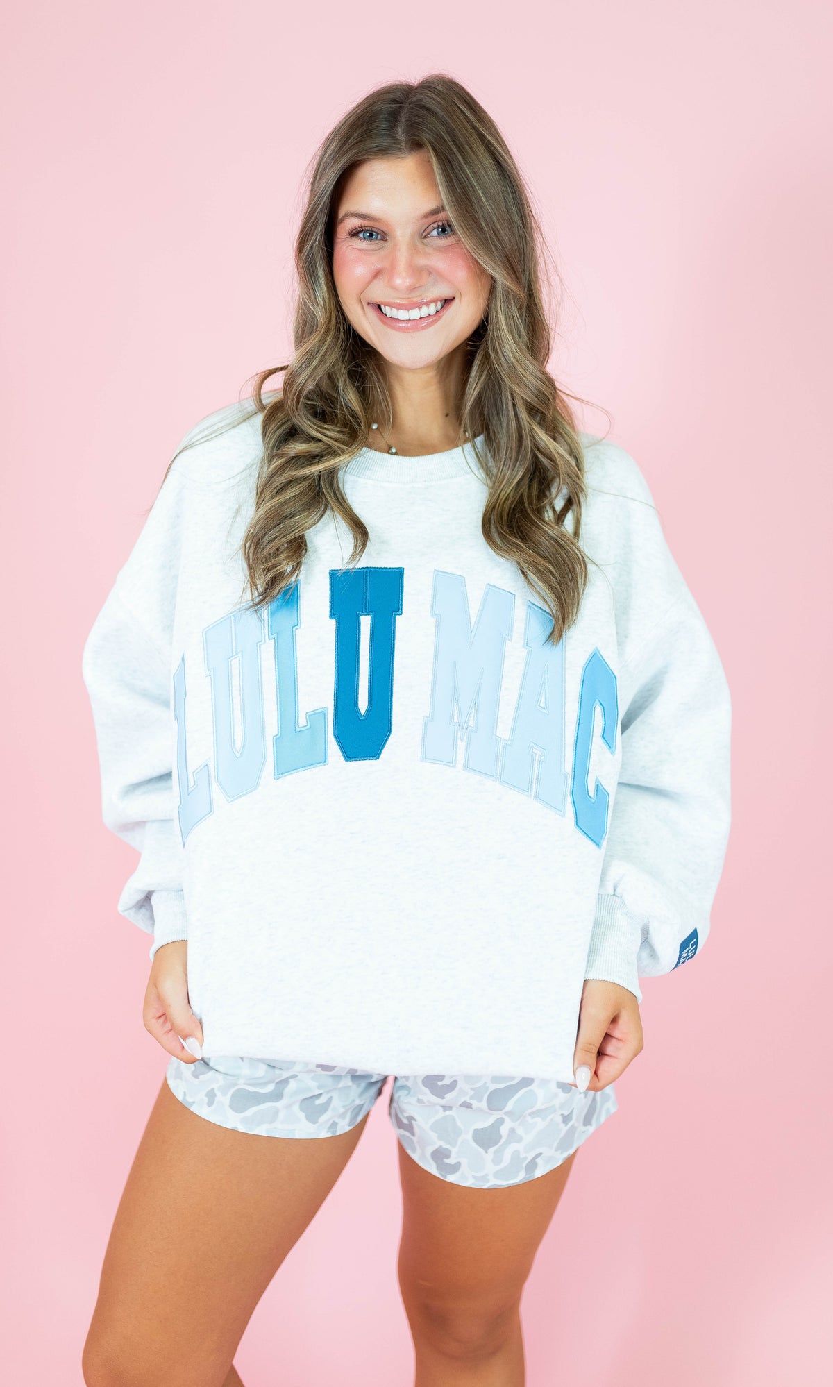 A smiling woman stands against a solid pink studio background wearing a white oversized sweatshirt with large light blue 'LOLU MAC' lettering across the front and matching patterned shorts.