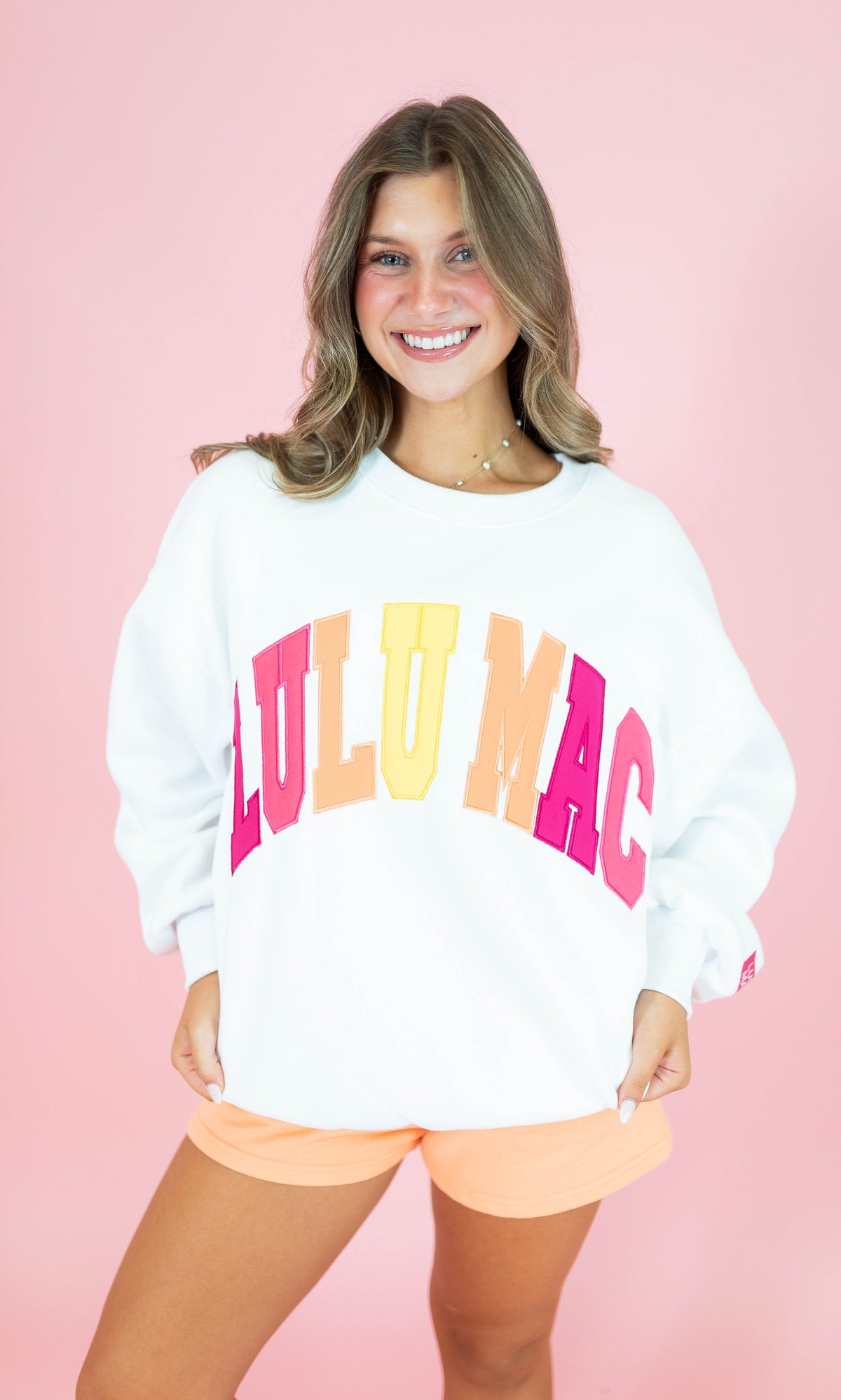 A smiling woman wearing a white crewneck sweatshirt with 'LULU MAC' printed across the front in large pink, orange, and yellow letters, paired with peach shorts, set against a solid pink background.