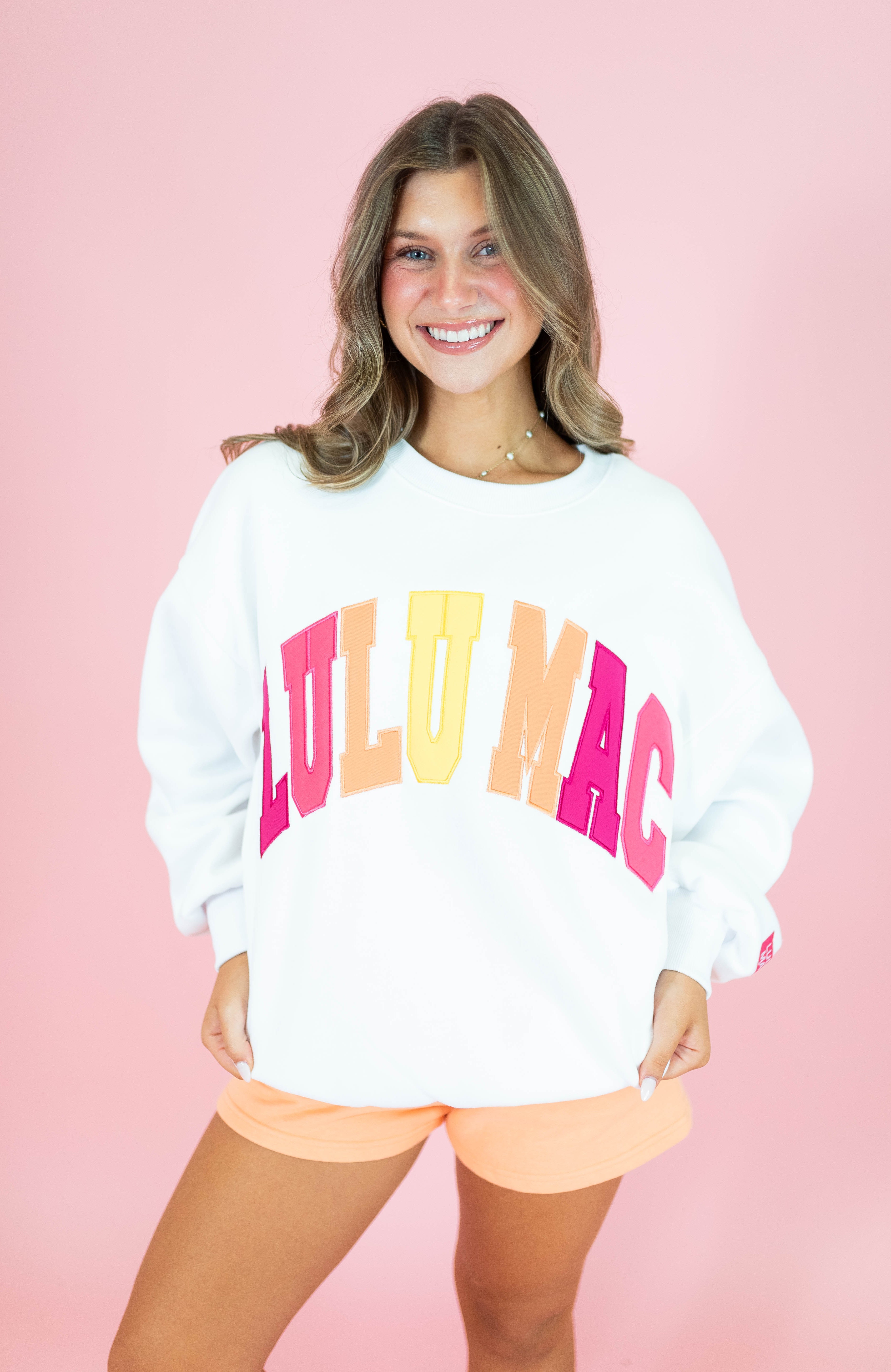 A smiling woman wearing a white crewneck sweatshirt with 'LULU MAC' printed across the front in large pink, orange, and yellow letters, paired with peach shorts, set against a solid pink background.