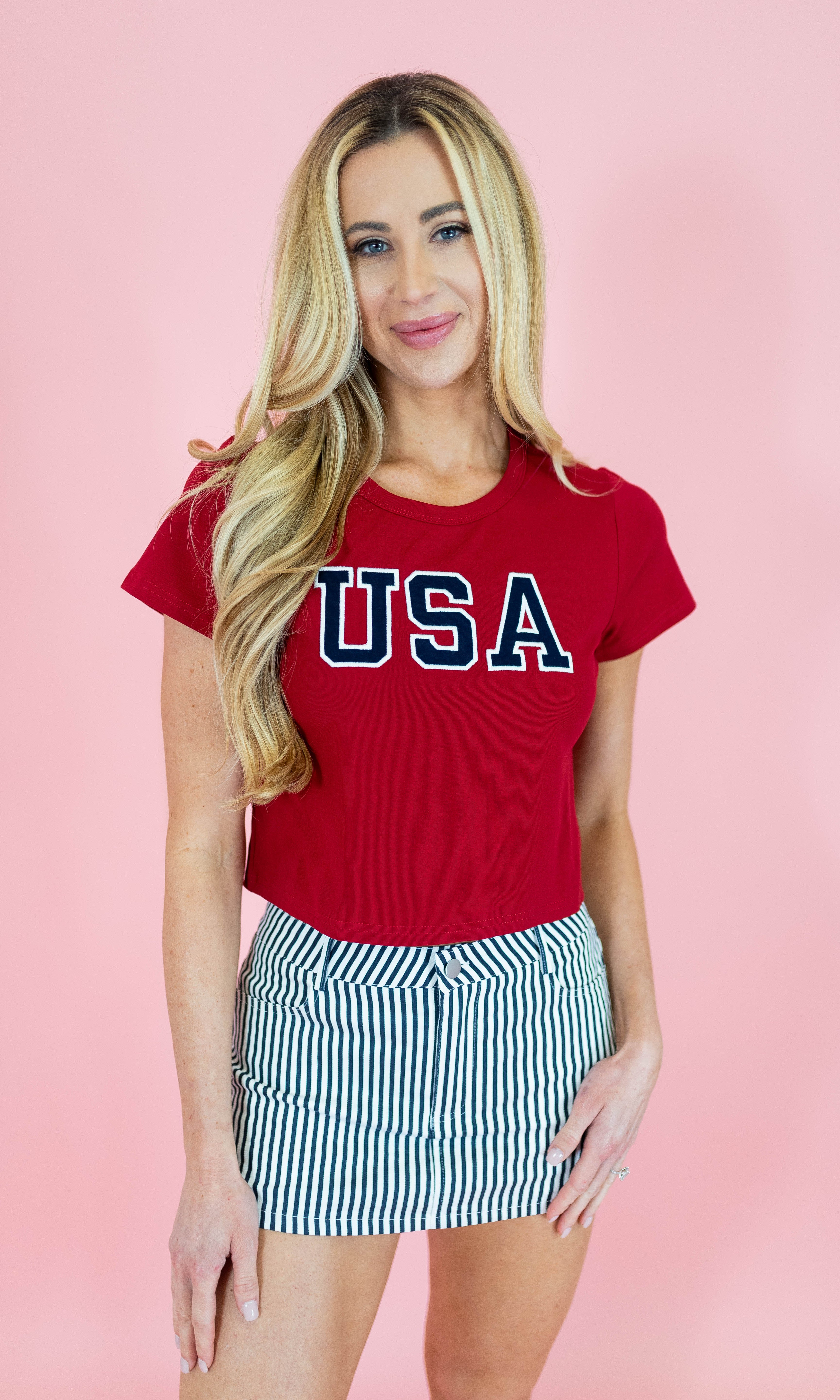 Woman wearing a red 'USA' t-shirt and striped skirt on a pink background