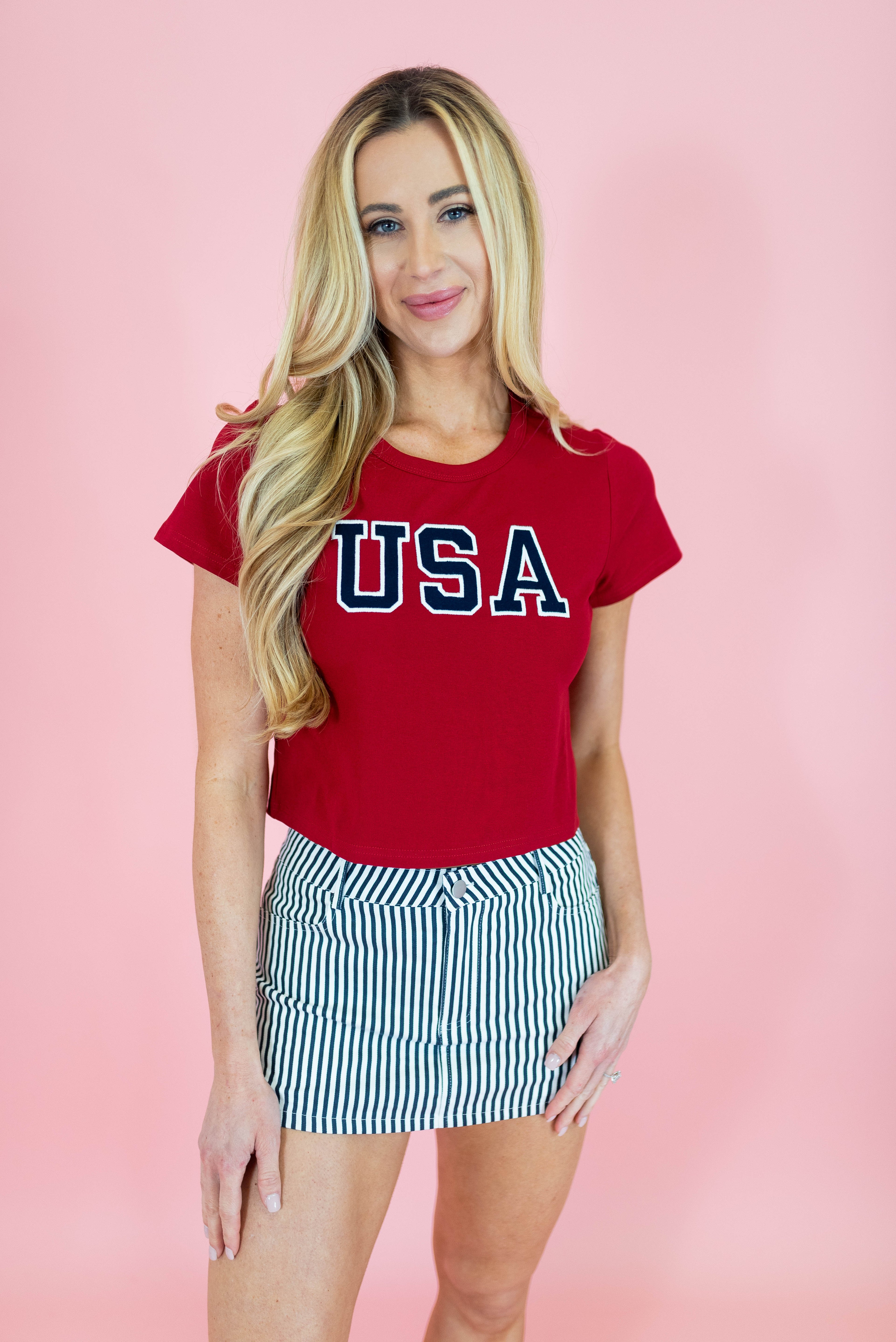 Woman wearing a red 'USA' t-shirt and striped skirt on a pink background