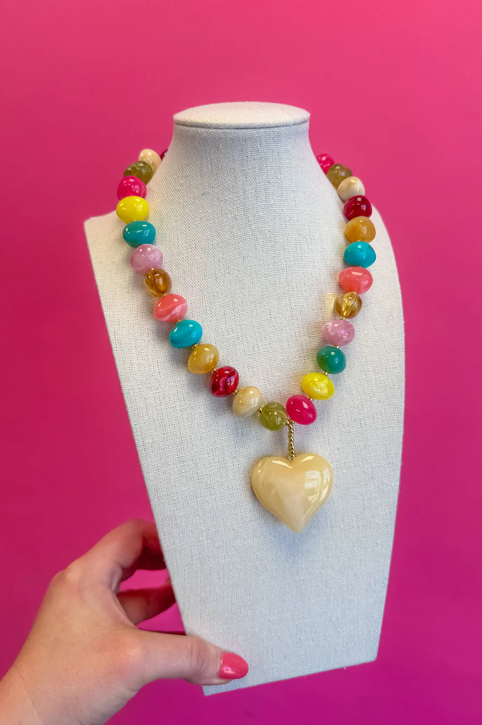 Colorful beaded necklace with a heart-shaped pendant on a mannequin bust against a pink background.
