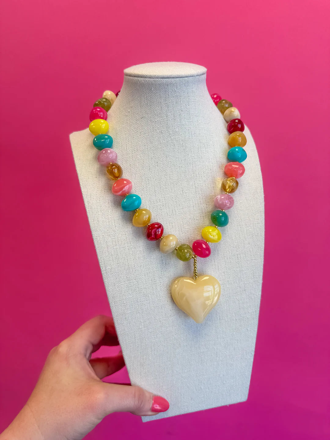 Colorful beaded necklace with a heart-shaped pendant on a mannequin bust against a pink background.