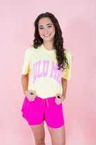 Person wearing a yellow t-shirt with pink text and pink shorts on a pink background
