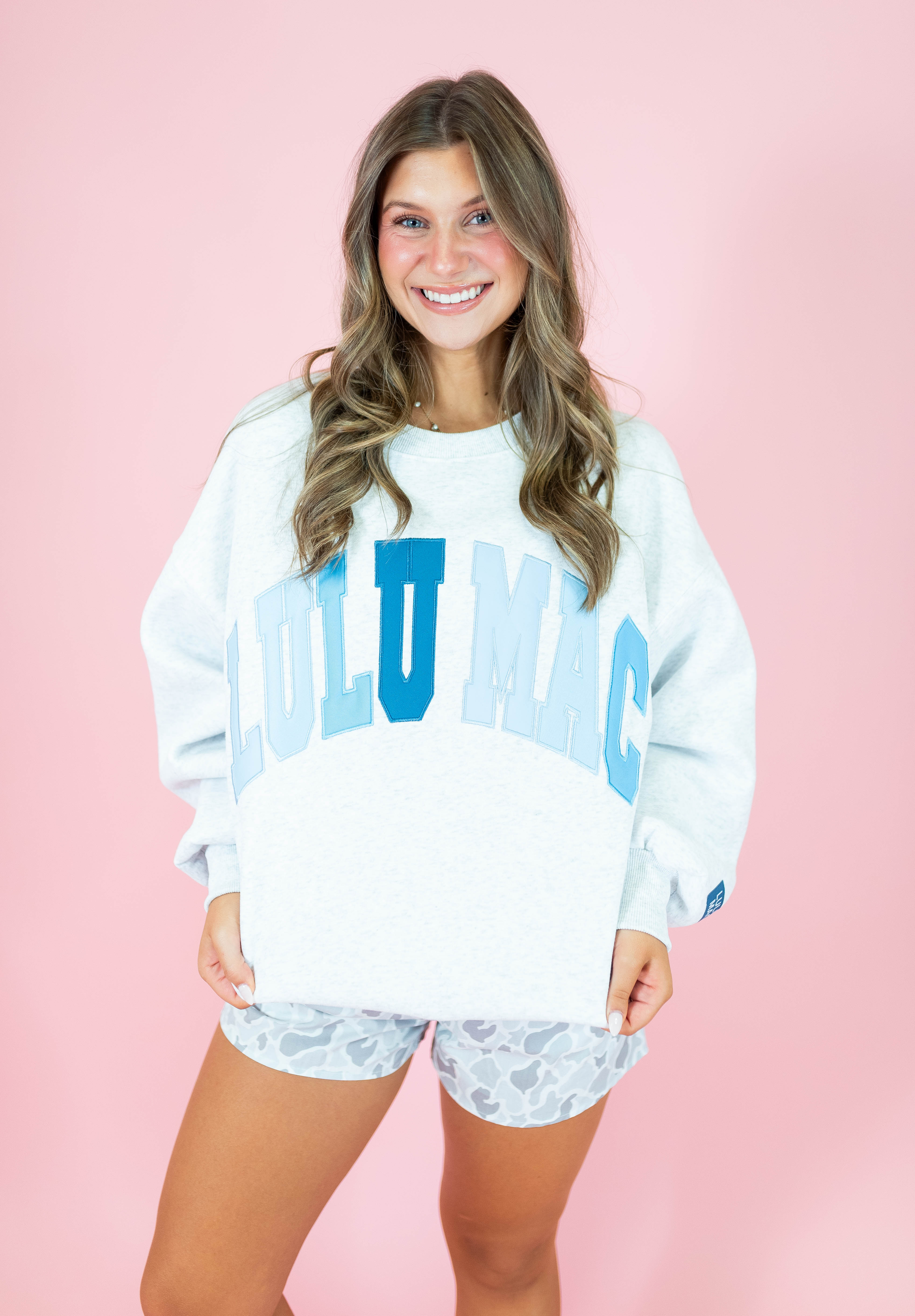 A smiling woman stands against a solid pink studio background wearing a white oversized sweatshirt with large light blue 'LOLU MAC' lettering across the front and matching patterned shorts.