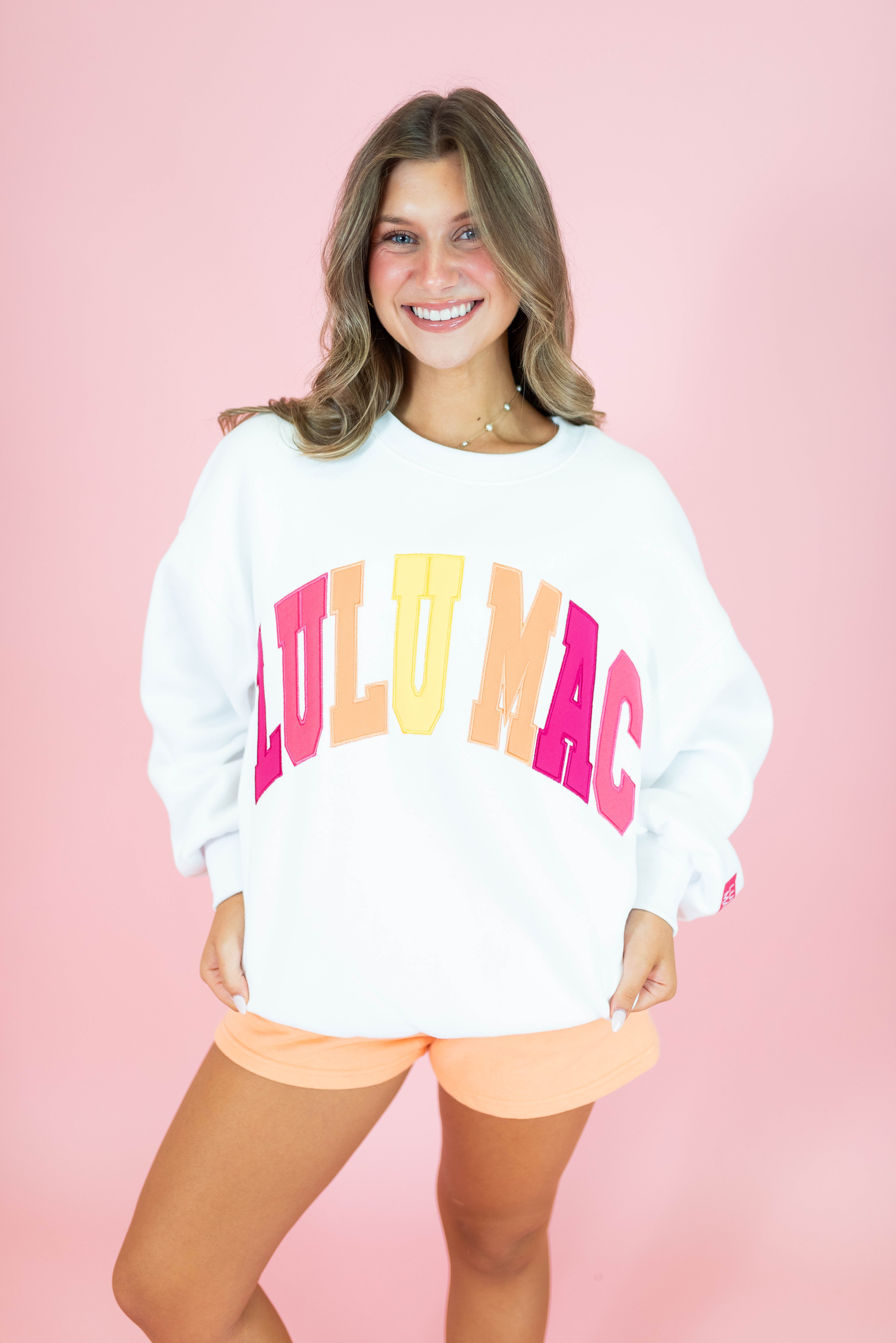 A smiling woman wearing a white crewneck sweatshirt with 'LULU MAC' printed across the front in large pink, orange, and yellow letters, paired with peach shorts, set against a solid pink background.