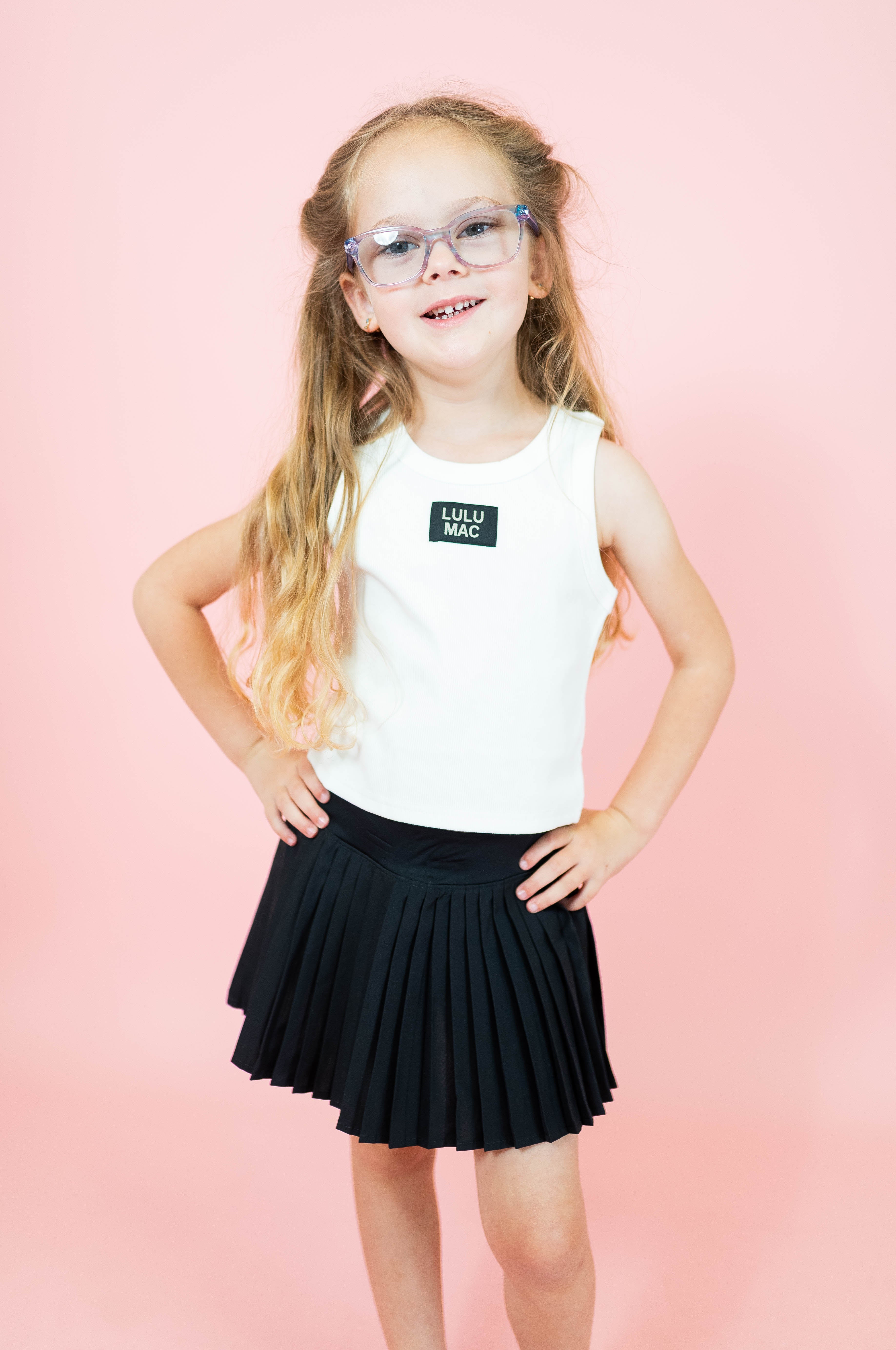 Young girl wearing a white t-shirt with a logo and a black pleated skirt on a pink background