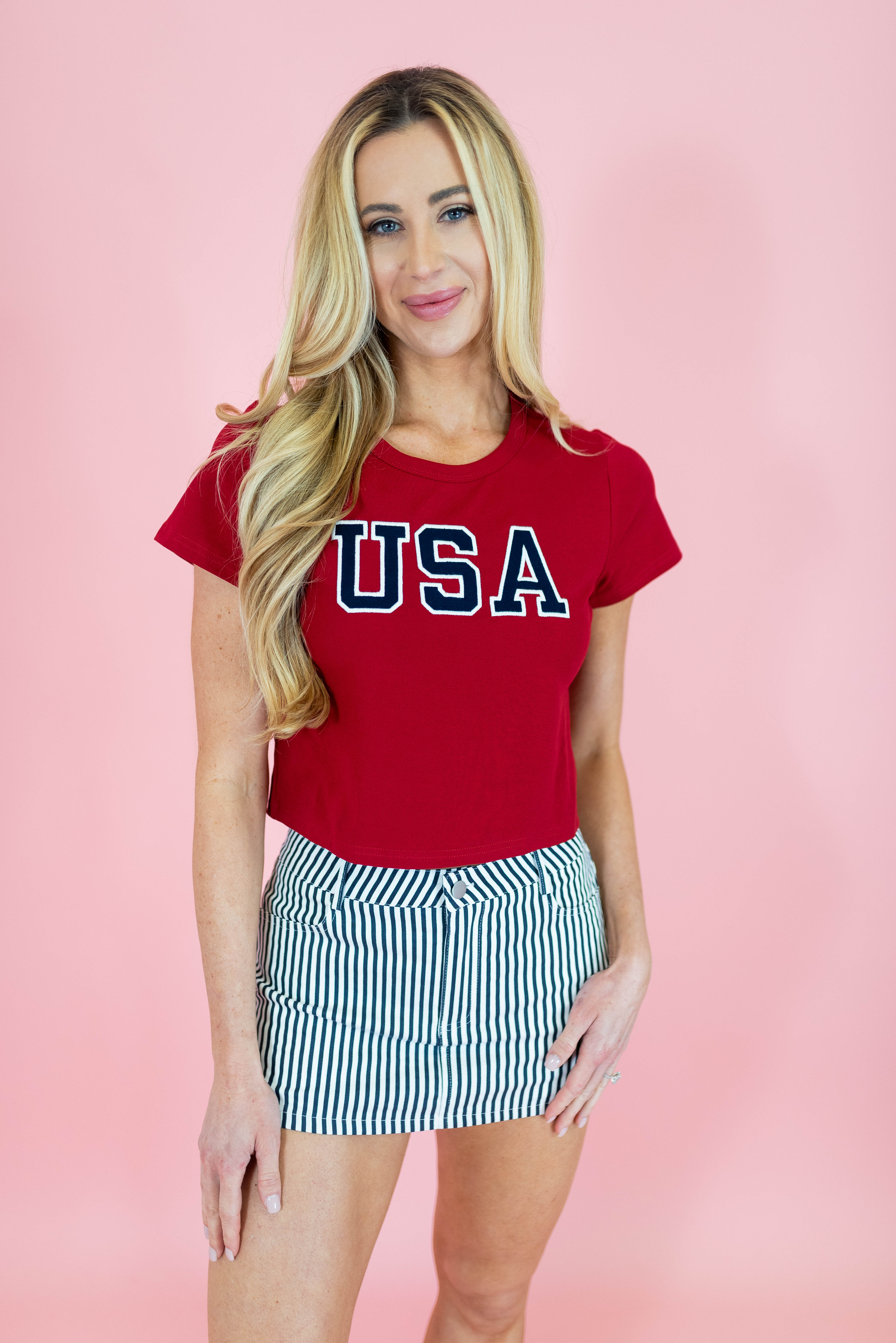 Woman wearing a red 'USA' t-shirt and striped skirt on a pink background
