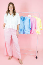Woman in white blouse and pink pants standing next to a rack with colorful shirts on a pink background