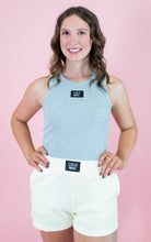 Woman wearing a light blue tank top and white shorts with visible branding against a pink background