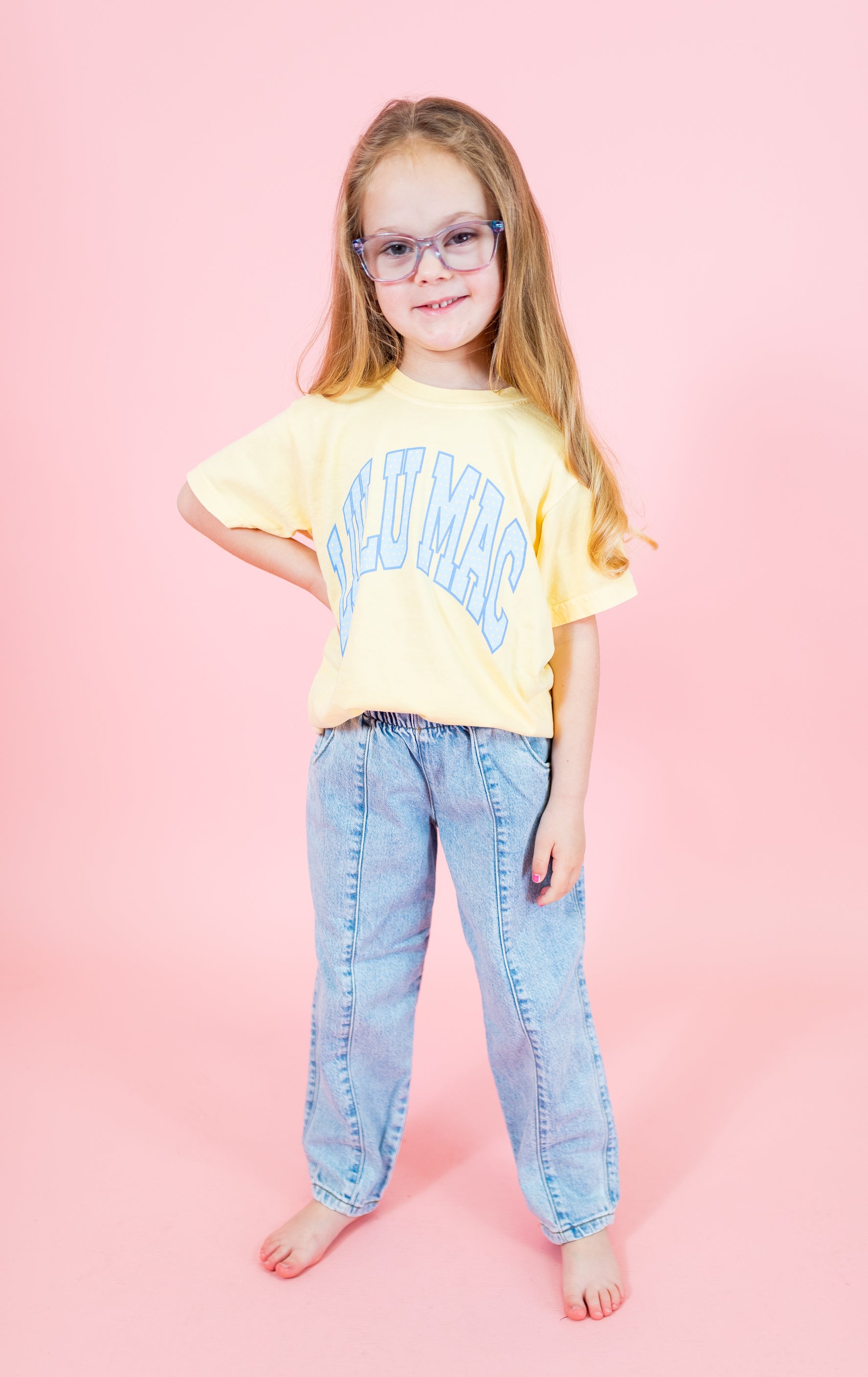 Young girl wearing a yellow t-shirt with text and blue jeans on a pink background