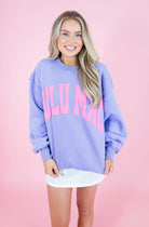 A smiling blonde woman wearing an oversized lavender crewneck sweatshirt with 'ULU MAC' printed in pink across the chest, paired with a white mini skirt, set against a solid pink studio background.