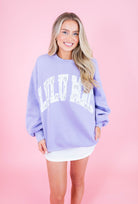 A woman modeling a lavender crewneck sweatshirt with 'LULU LULU' printed in white arched letters across the chest, paired with a white mini skirt, set against a solid pink studio background.